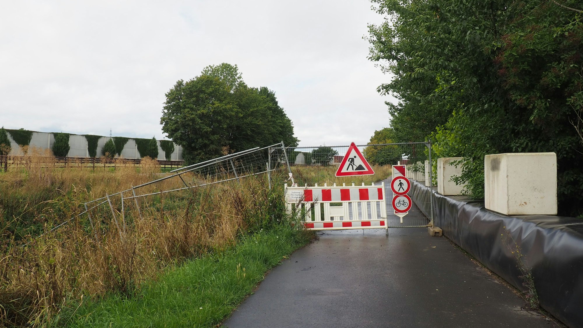 Ein asphaltierter Weg ist mit Baken und Bauzaun-Elementen versperrt.