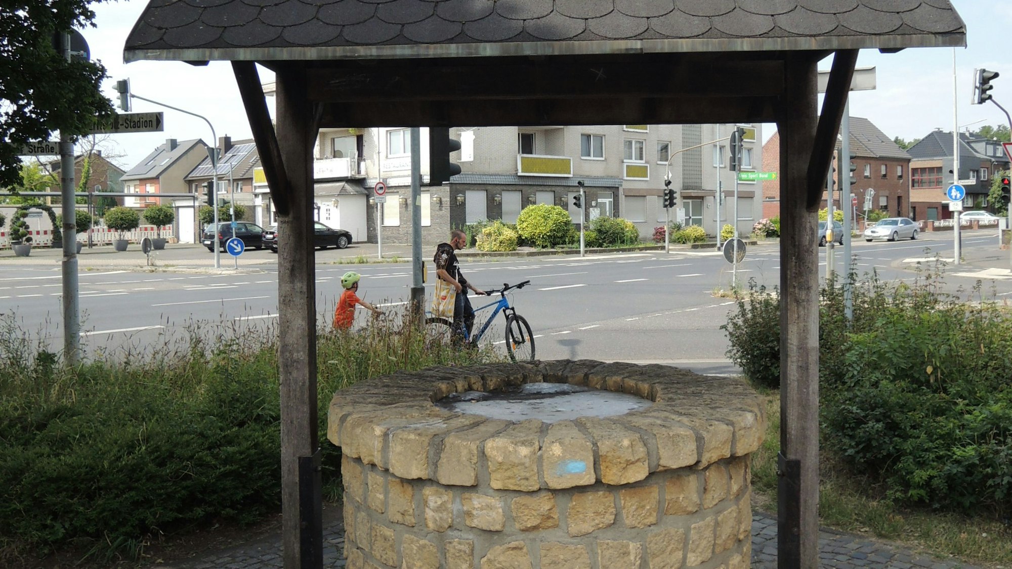 Ein überdachter Brunnen aus Steinen steht an einer Straße.