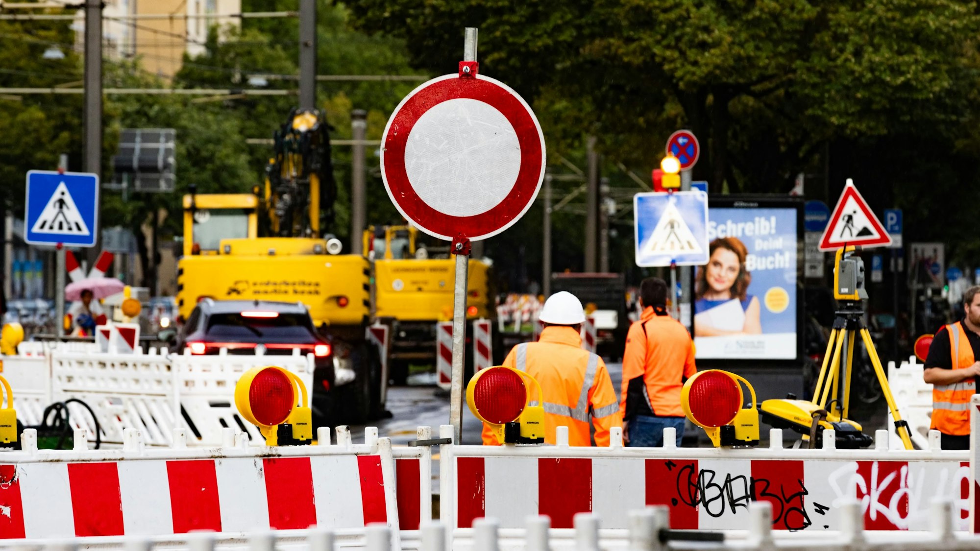 Hinter einem Baustellenzaun stehen Arbeiter und zahlreiche Straßenschilder.