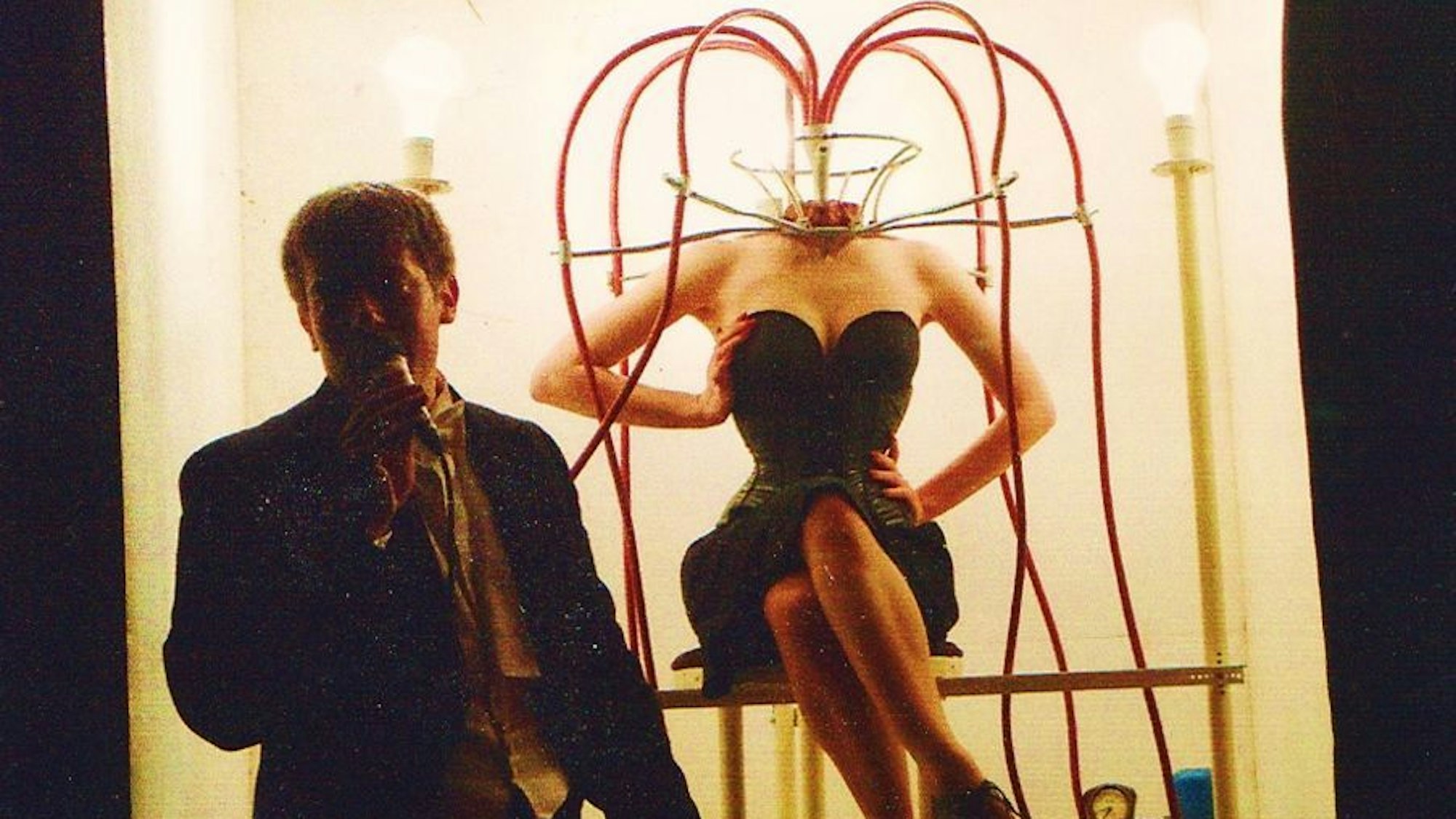 Ein Mann im Frack steht vor einem erhellten Podest, auf dem eine knapp bekleidete Frau ohne Kopf sitzt. An ihrem Hals laufen Schläuche zusammen.