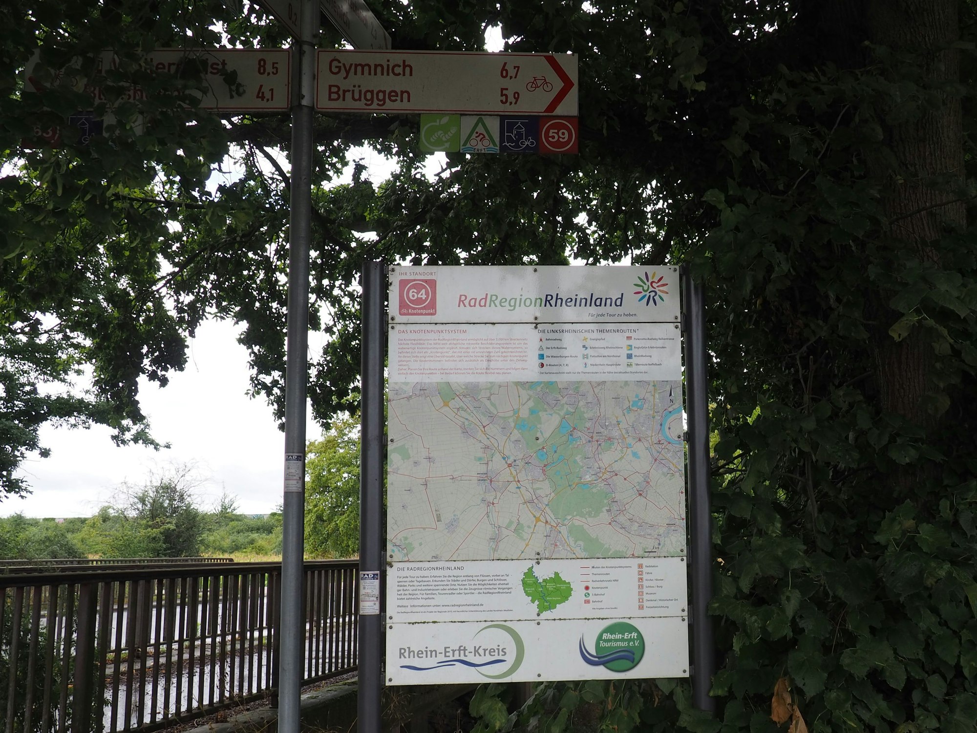 Radweg-Schilder zeigen nach Gymnich und Brüggen, daneben hängt eine Karte.