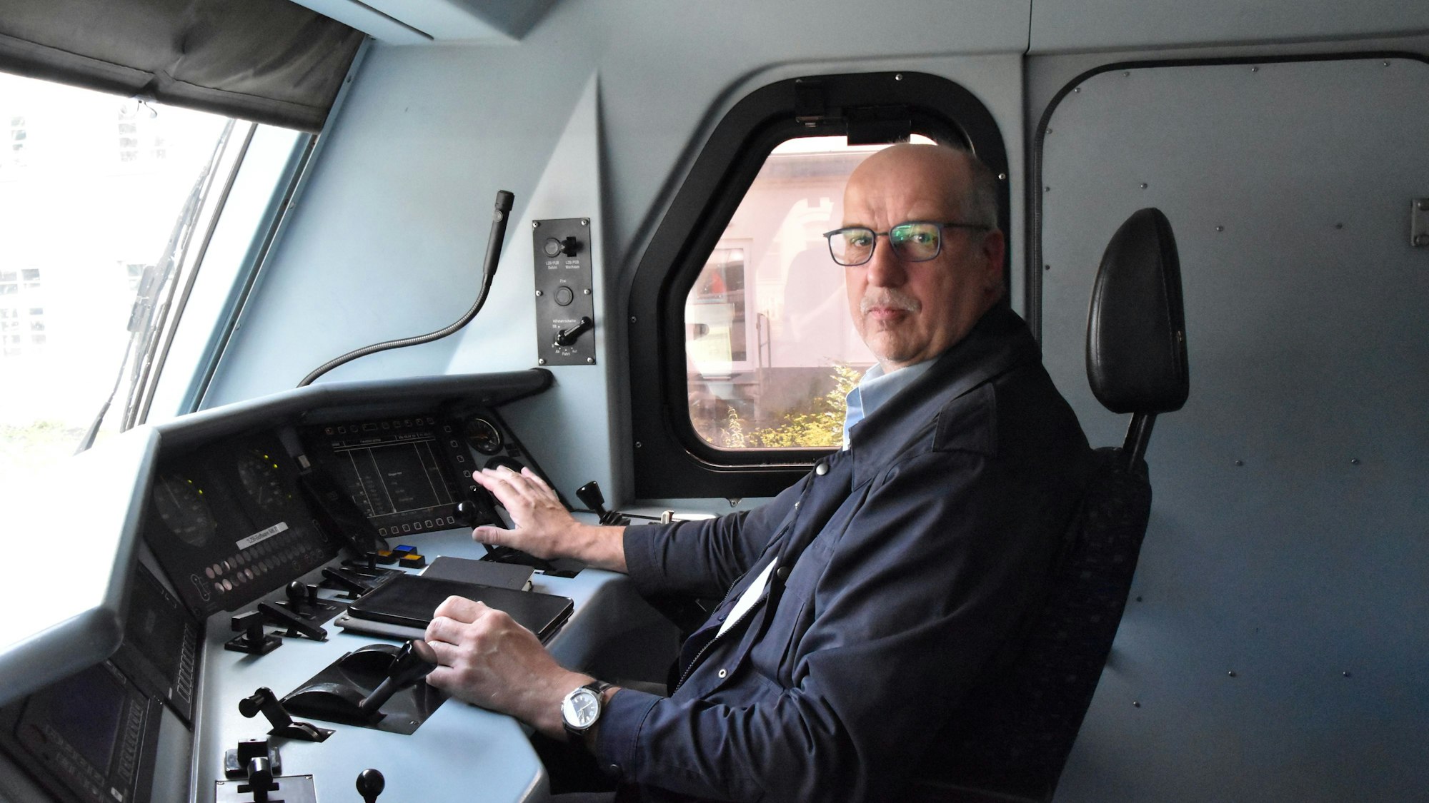 Ein Mann mit Brille sitzt im Führerstand einer Eisenbahn-Lokomotive. Er trägt eine blaue Jacke; vor sich hat er das Armaturenbrett der Lok.
