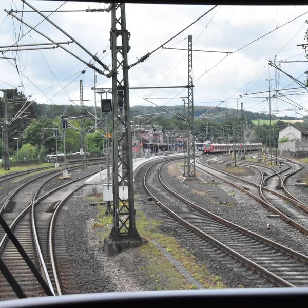 Blick aus dem Führerstand eines Regionalexpresszugs. Zu sehen sind die Schienen, Oberleitungsmasten und ein rotes Bahnhofsgebäude im Hintergrund. Auf einem Gleis stehen rote Waggons einer S-Bahn.