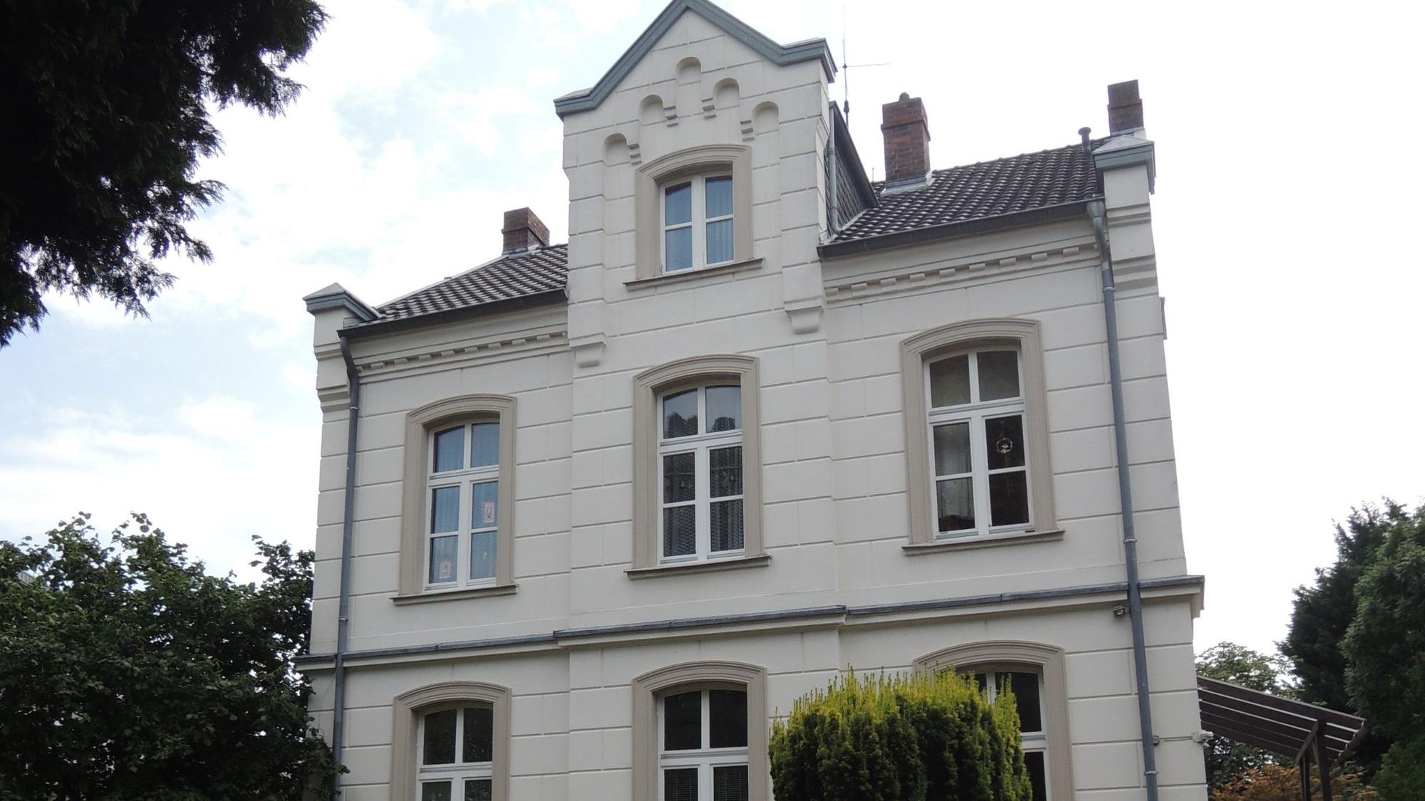 Ein Gebäude aus hellem Stein steht an einer Straße, drum herum sind Büsche und Bäume.