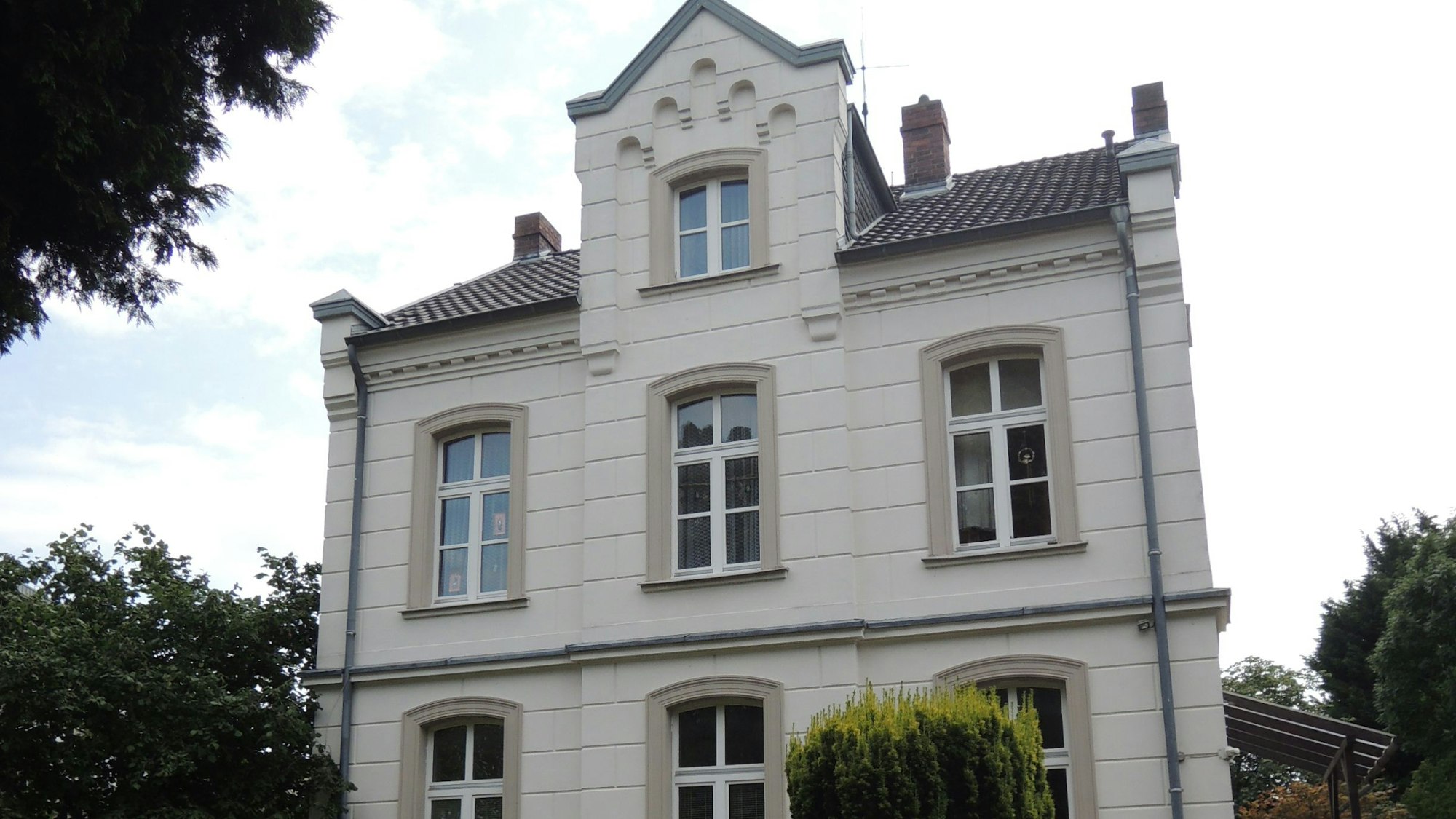 Ein Gebäude aus hellem Stein steht an einer Straße, drum herum sind Büsche und Bäume.