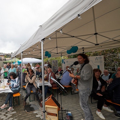 Stephan Brings begleitet sich beim Singen selbst mit der Gitarre. Die Gäste der Einweihungsfeier hören zu, im Hintergrund das neu gebaute Haus
