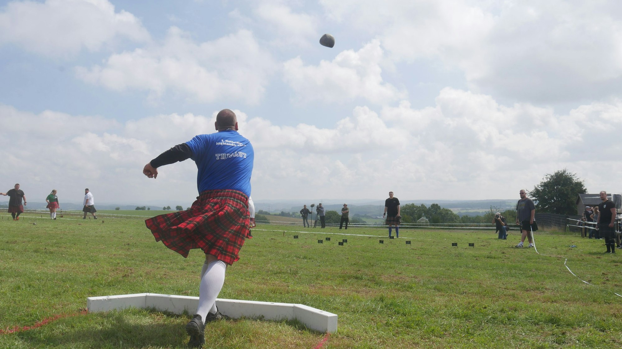 Ein Mann im Schottenrock stößt einen Stein über eine Wiese.