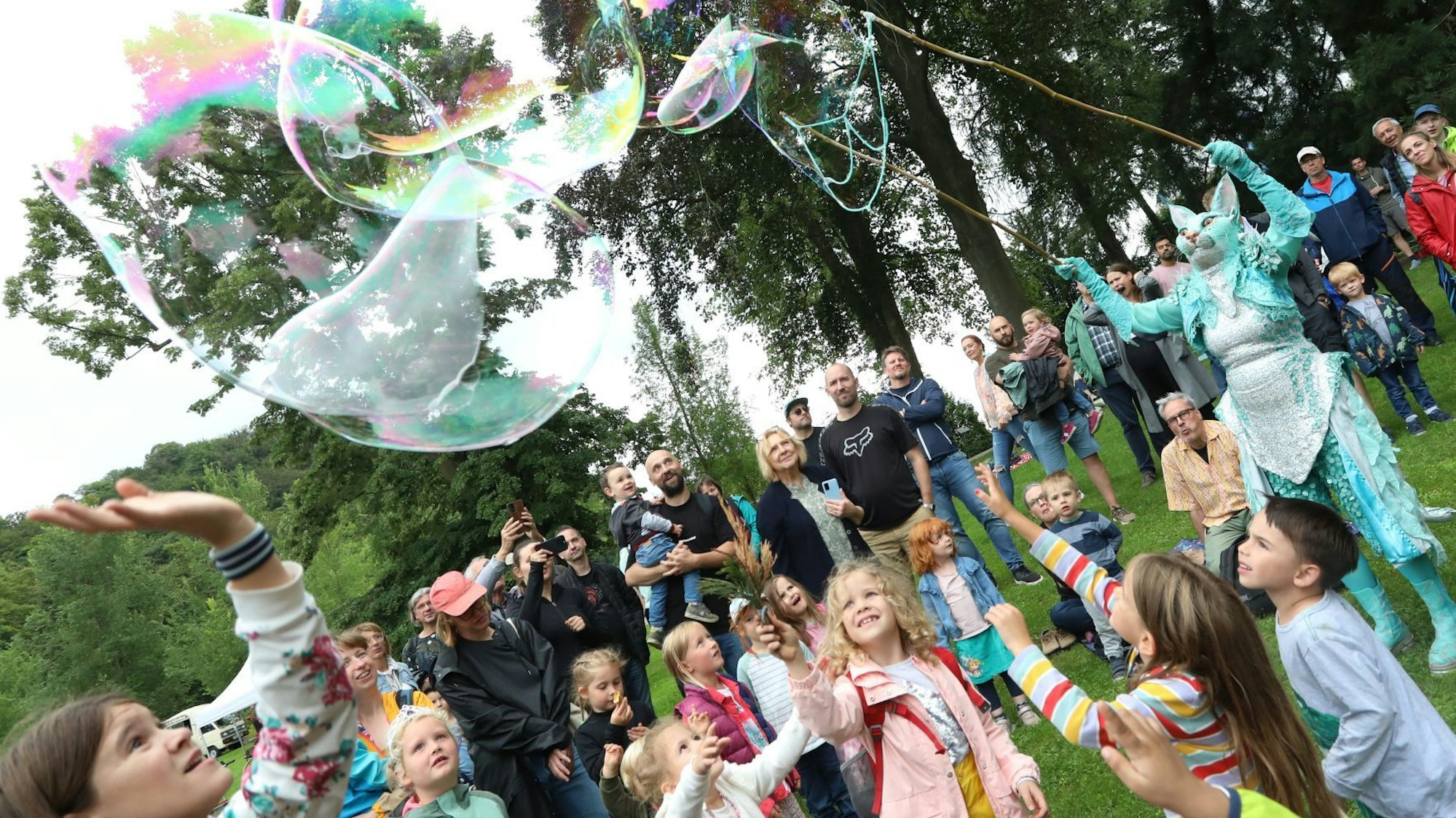 Spielende Kinder versuchen große Seifenblasen zum Zerplatzen zu bringen, die eine Darstellerin im blauen Katzenkostüm produziert hat.