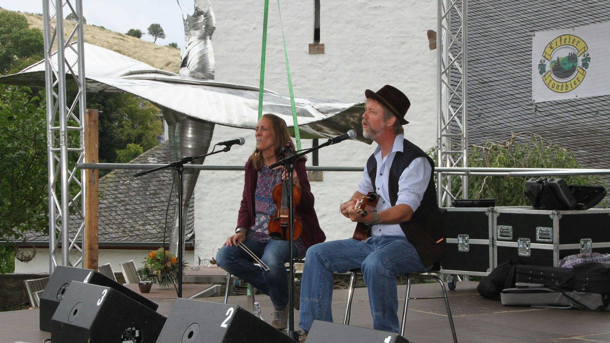 Das Bild zeigt das Musiker-Duo Yopi und Lotta, das vor einer Stahlblech-Skulptur musiziert.
