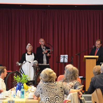 Eine Frau in Schwesterntracht mit Fahrrad und ein Mann mit einem Autolenkrad in der Hand stehen neben dem Vortragenden am Pult.