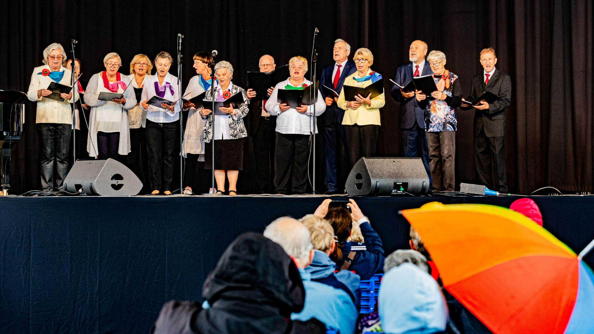 Ein Chor steht auf der Bühne während Leute mit Regenschirmen im Publikum sitzen.