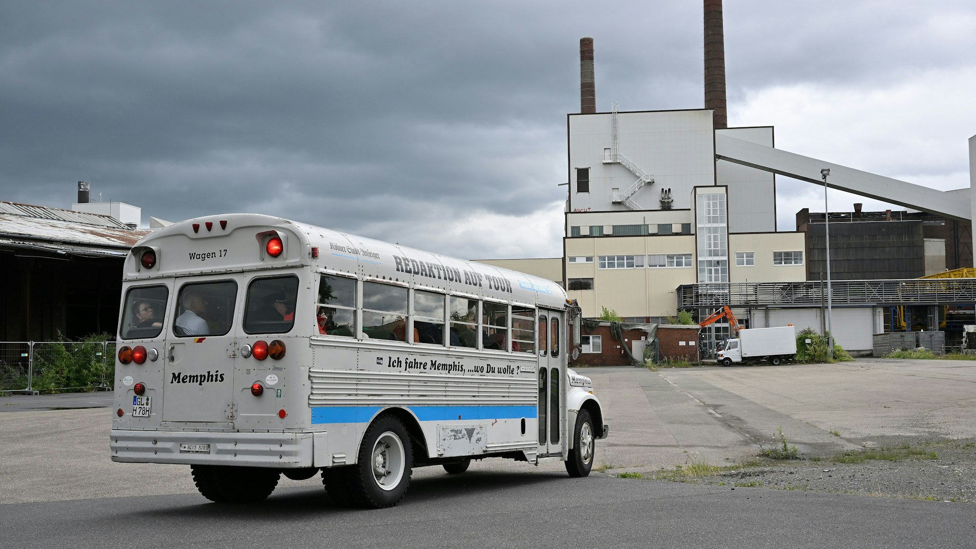 Ein historischer amerikanischer Schulbus mit der Aufschrift „Redaktion auf Tour“ fährt über das Bergisch Gladbacher Zanders-Gelände auf das dortige Kraftwerk mit Schornsteinen zu,