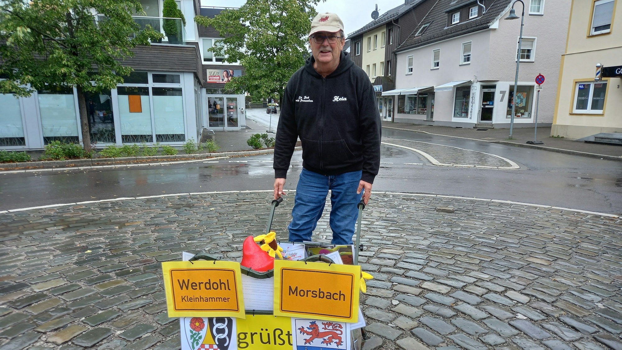 Reinhard Kaldeich steht mit einer mit den Ortsschildern von Werdohl und Morsbach geschmückten Schubkarre auf dem leeren Milly-la-Forêt-Platz in Morsbach.