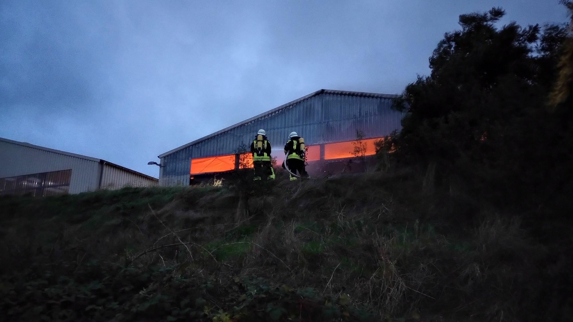 Man sieht zwei Feuerwehrleute auf einem Hang, im Hintergrund die Halle, aus der ein Leuchten kommt.
