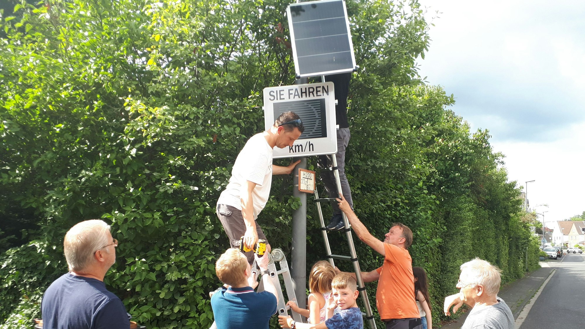 Mehrere freiwillige Helfer montierten mit vereinten Kräften die neue Tempo-Infotafel an der Longericher Straße. Im Hintergrund ist eine grüne Hecke zu sehen.