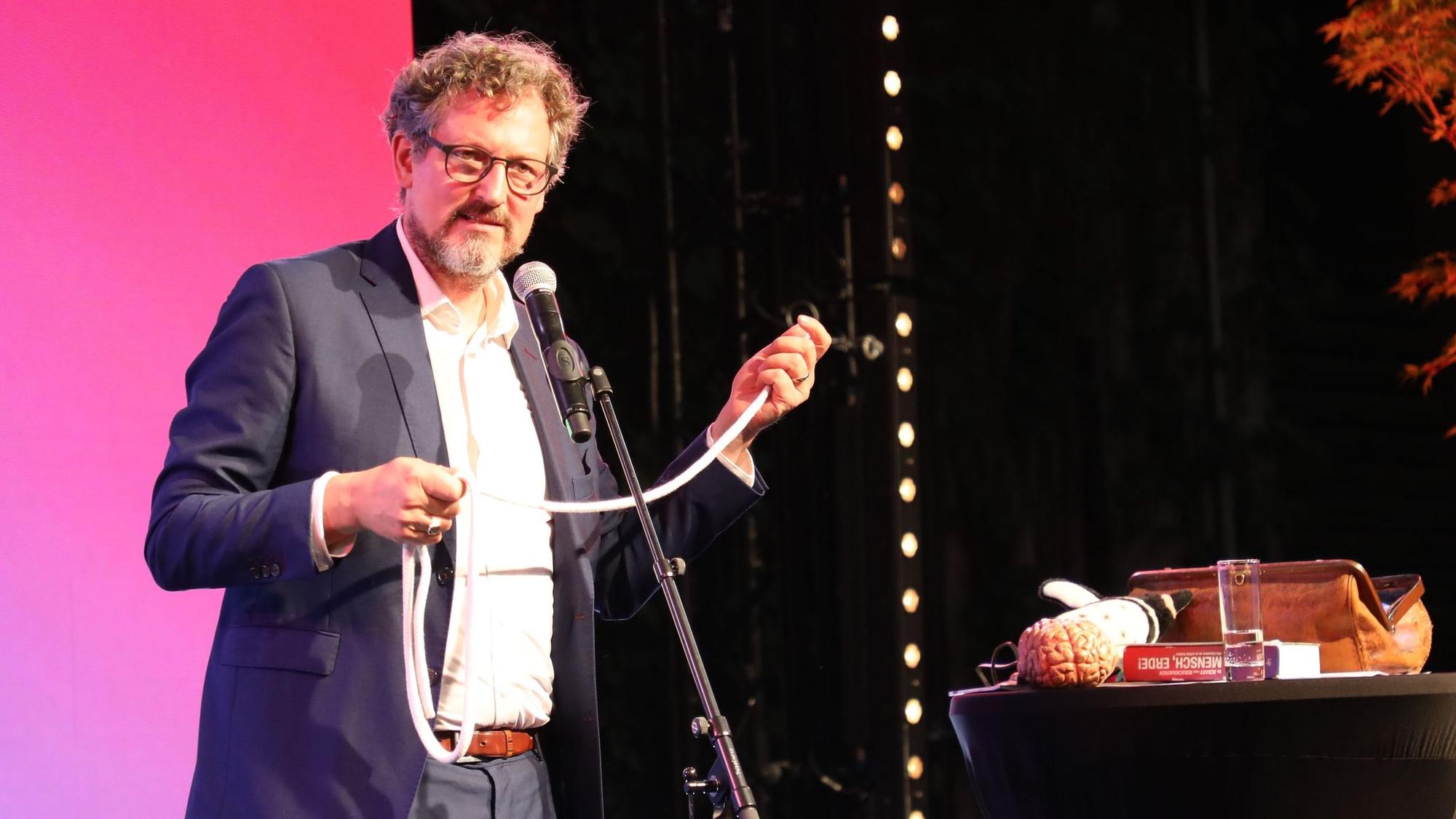 Dr. Eckart von Hirschhausen steht mit einem Seil in den Händen auf einer Bühne und spricht in ein Mokrofon.