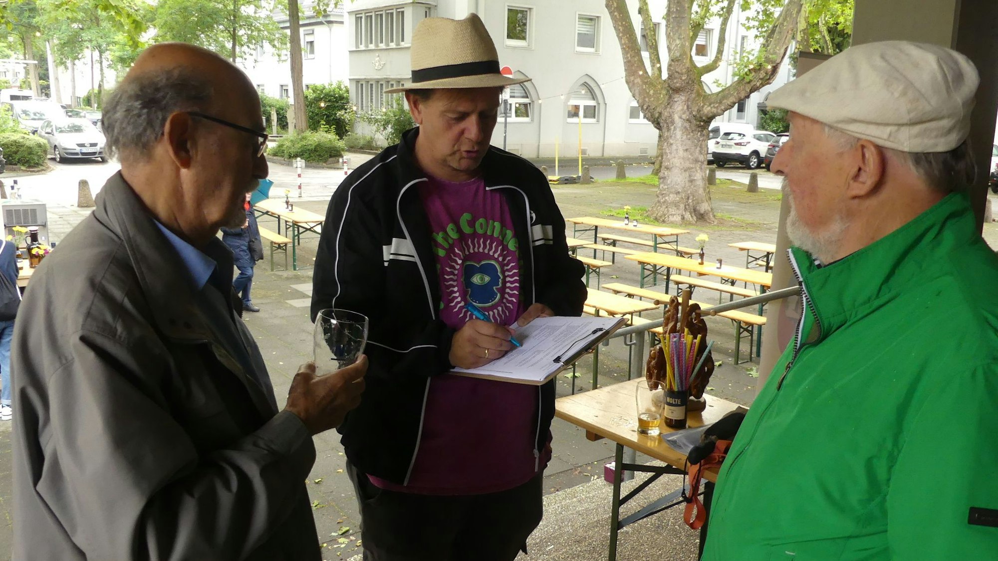 Ein Mann mit Strohhut notiert Wünsche auf einem Klemmbrett. Zwei Männer stehen links und rechts von ihm.