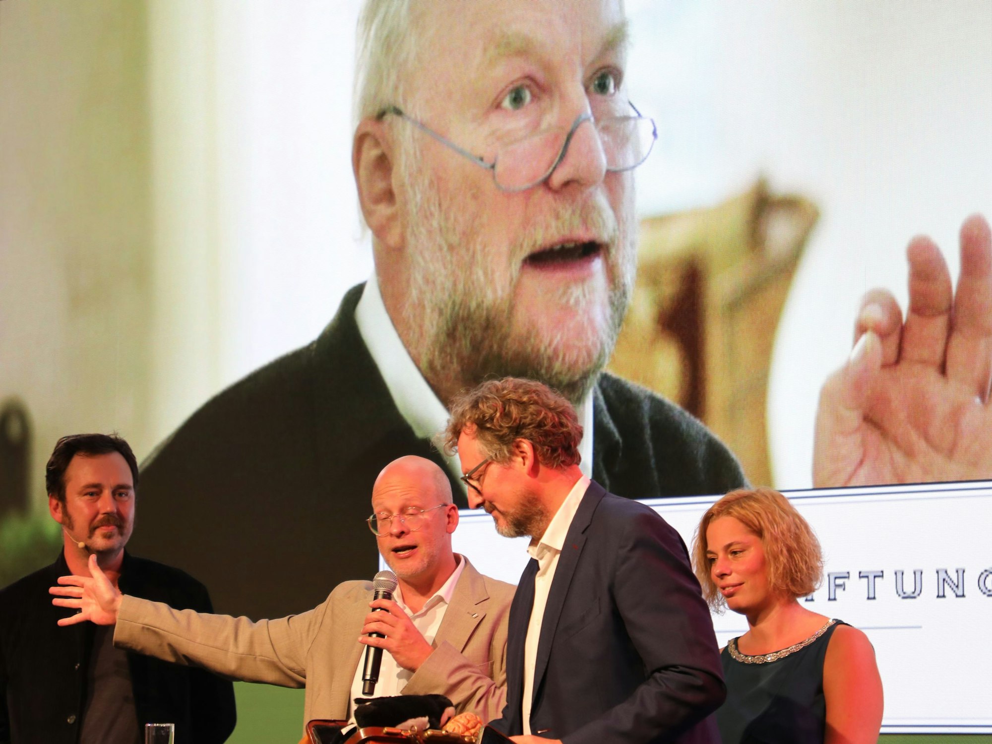 Dr. Eckhart von Hirschhausen steht mit Laudator Eric Wrede, Hanna Roth und Bruder David Rot bei der Preisverleihung des Fritz Roth-Medienpreises vor einer Leinwand, auf der ein großes Bild des verstorbenen Fritz Roth zu sehen ist,