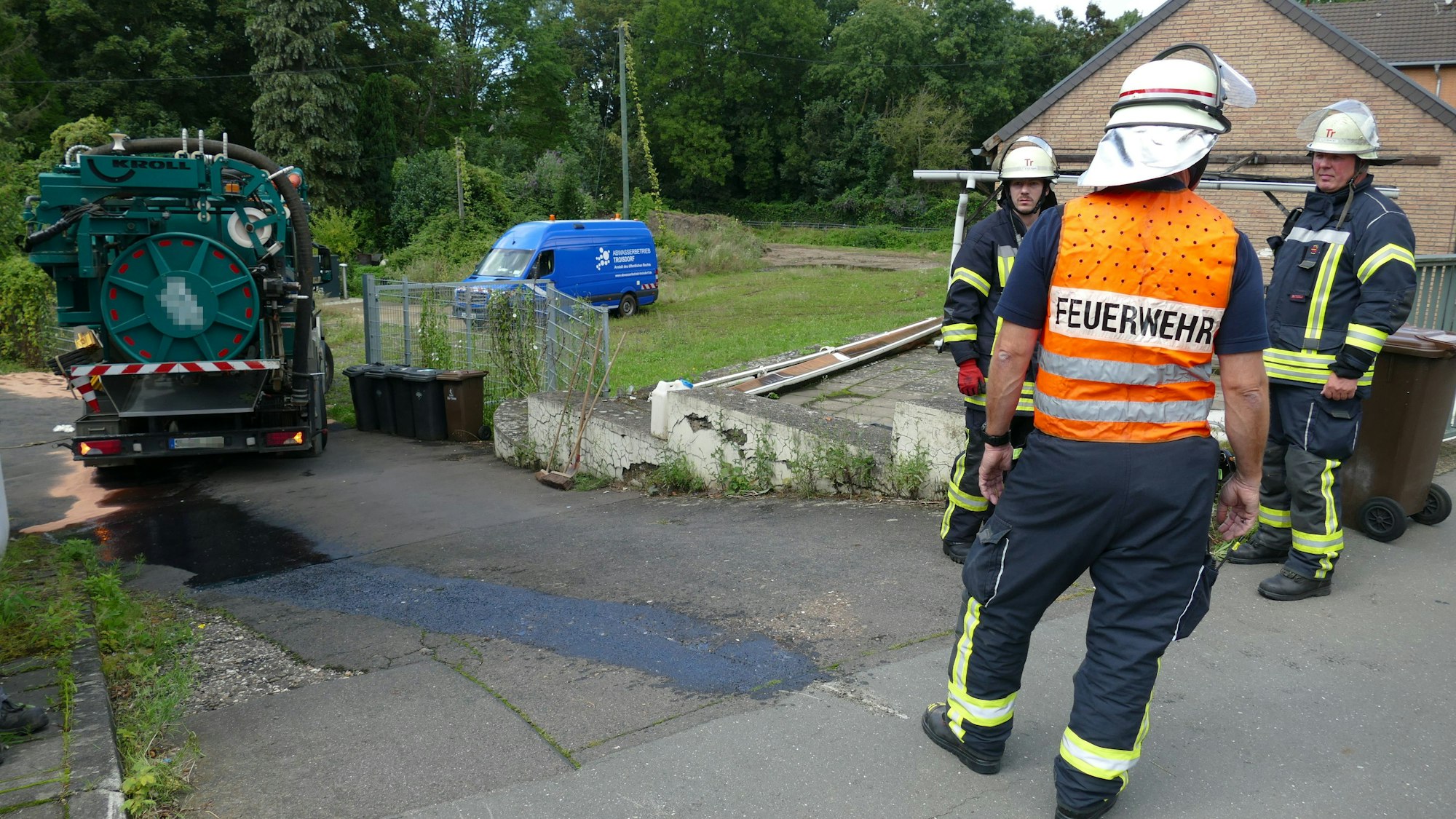 Drei Mitglieder der Feuerwehr stehen vor einer Ölspur. Ein Mann trägt eine Warnweste mit der Aufschrift Feuerwehr, im Hintergrund steht ein grünes Pumpfahrzeug, aus dessen Tank Öl gelaufen ist.