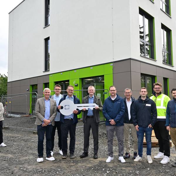 Vor dem Neubau der Modulschule steht der Bürgermeister mit den am Projekt beteiligten Fachleuten. Sie halten einen großen symbolischen Schlüssel in der Hand.