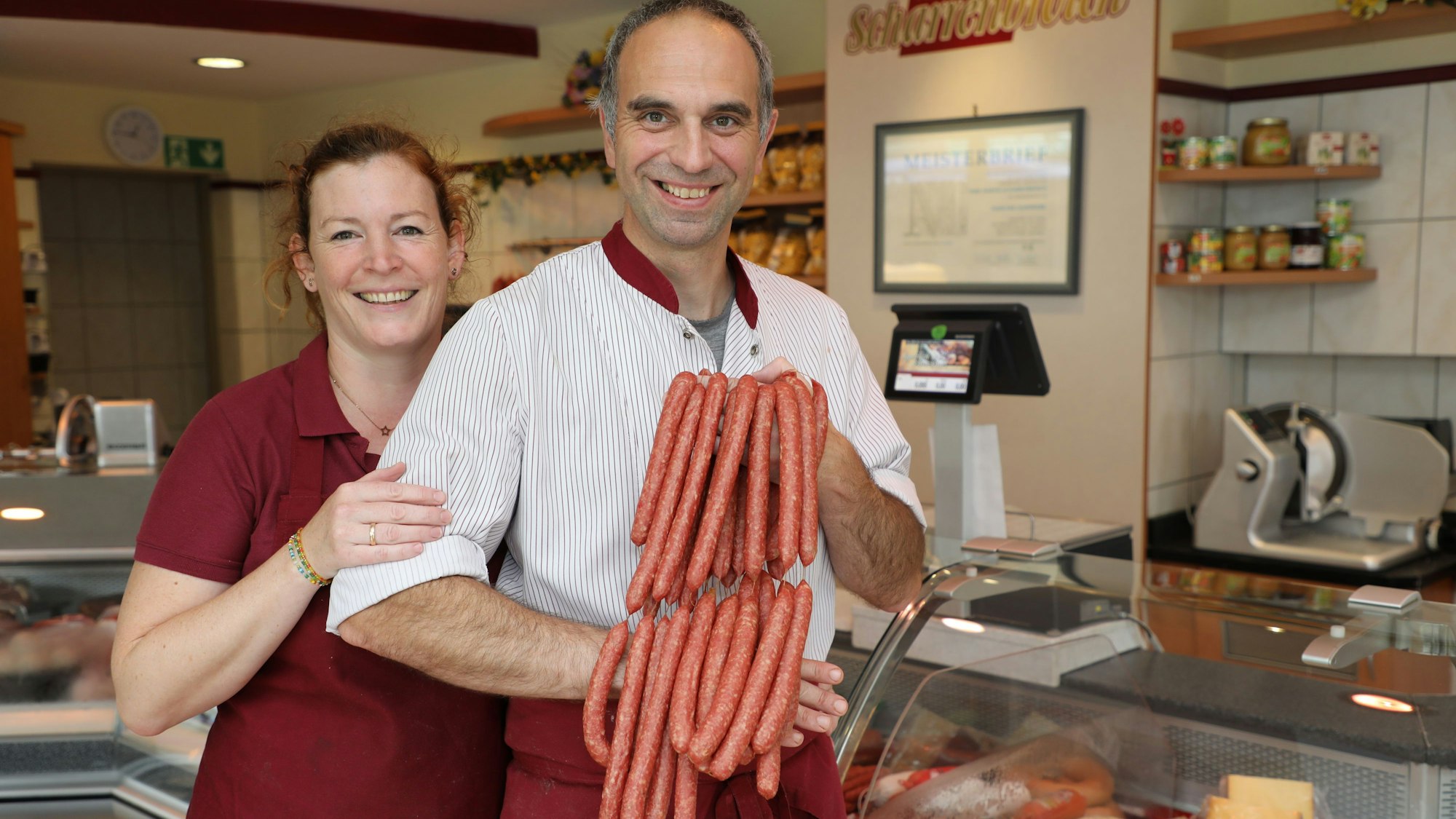 Metzger Martin Scharrenbroich hält Würste vor seiner Verkaufstheke in die Höhe, neben ihm steht seine Frau Nicola Scharrenbroich.