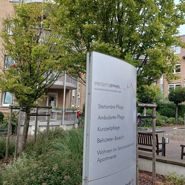 Ein großes Schild vor dem Seniorenzentrum weist auf die unterschiedlichen Abteilungen hin. Im Hintergrund stehen Bänke und Bäume.