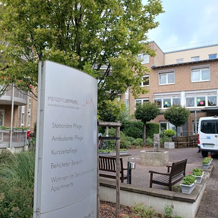 Ein großes Schild vor dem Seniorenzentrum weist auf die unterschiedlichen Abteilungen hin. Im Hintergrund stehen Bänke und Bäume.