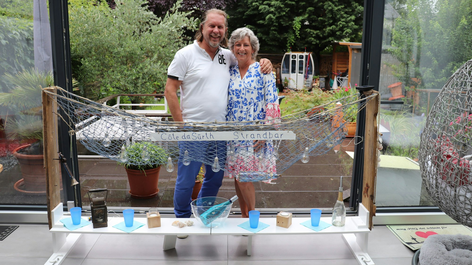 Ein Paar steht hinter einem selbstgebauten Regal mit einem Fischernetz, einer Lichterkette und allerlei Deko.