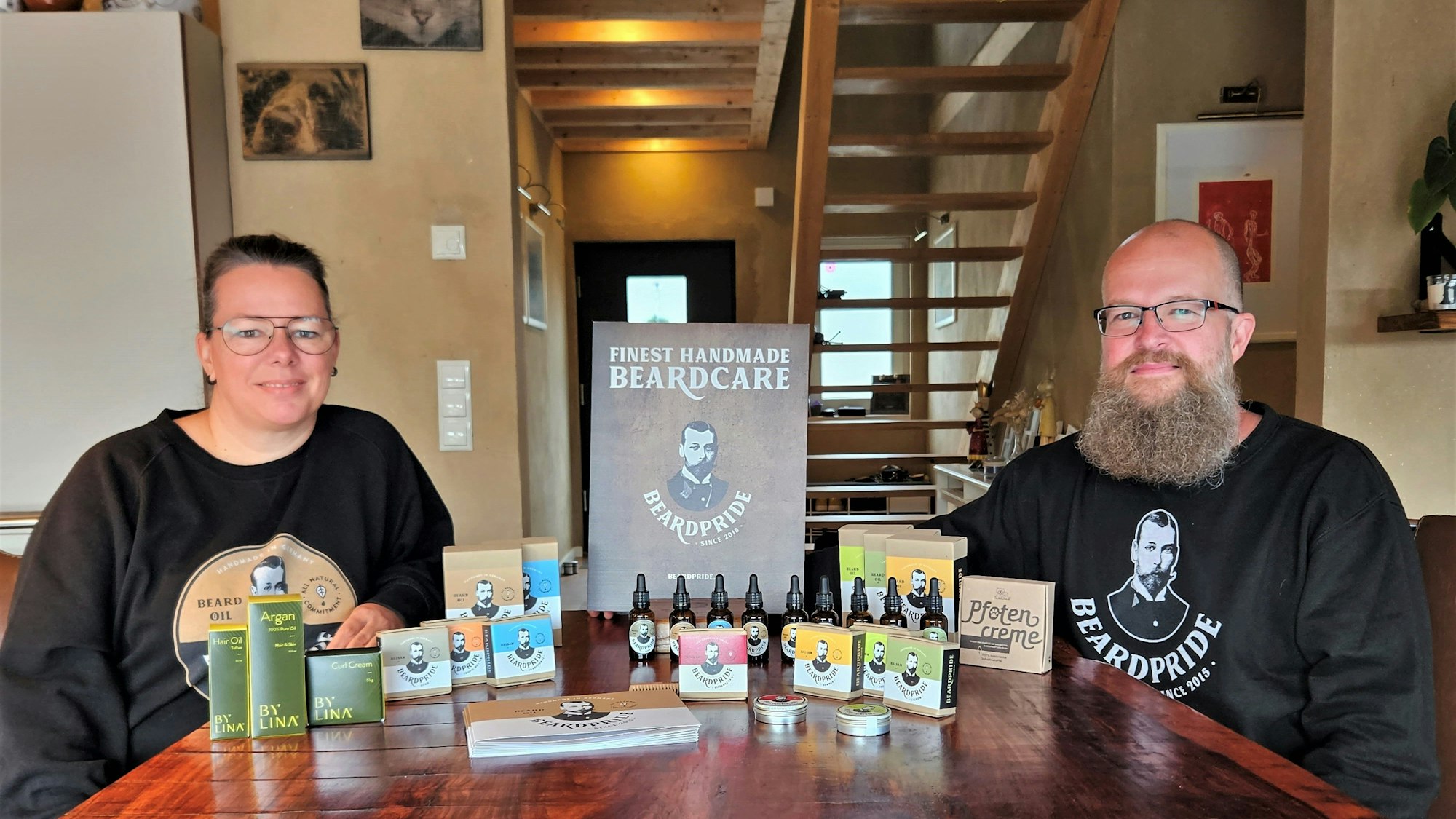 Miriam und Marko Schulze sitzen an einem Tisch, auf dem Öle und Balsame für die Bartpflege zu sehen sind.