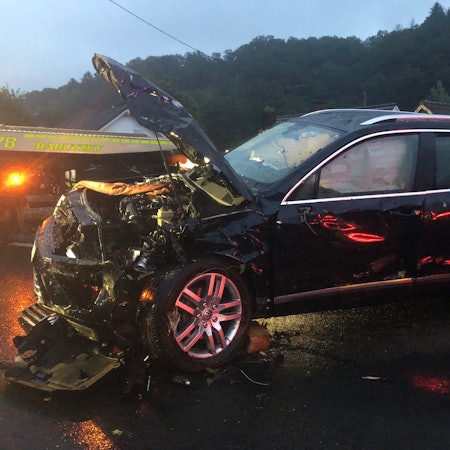 Ein schwer beschädigtes Auto steht auf einer Straße in der Dämmerung, im Hintergrund steht ein Abschleppfahrzeug.