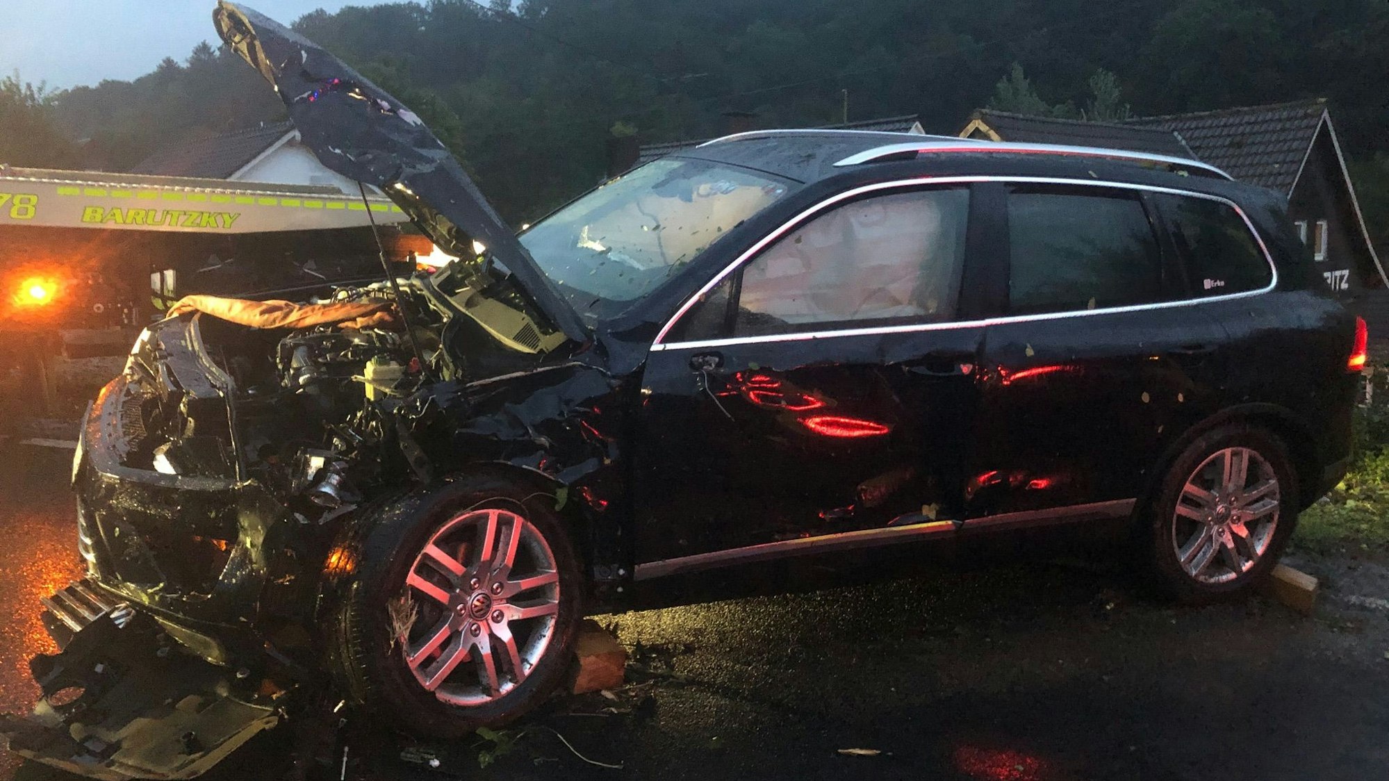 Ein schwer beschädigtes Auto steht auf einer Straße in der Dämmerung, im Hintergrund steht ein Abschleppfahrzeug.