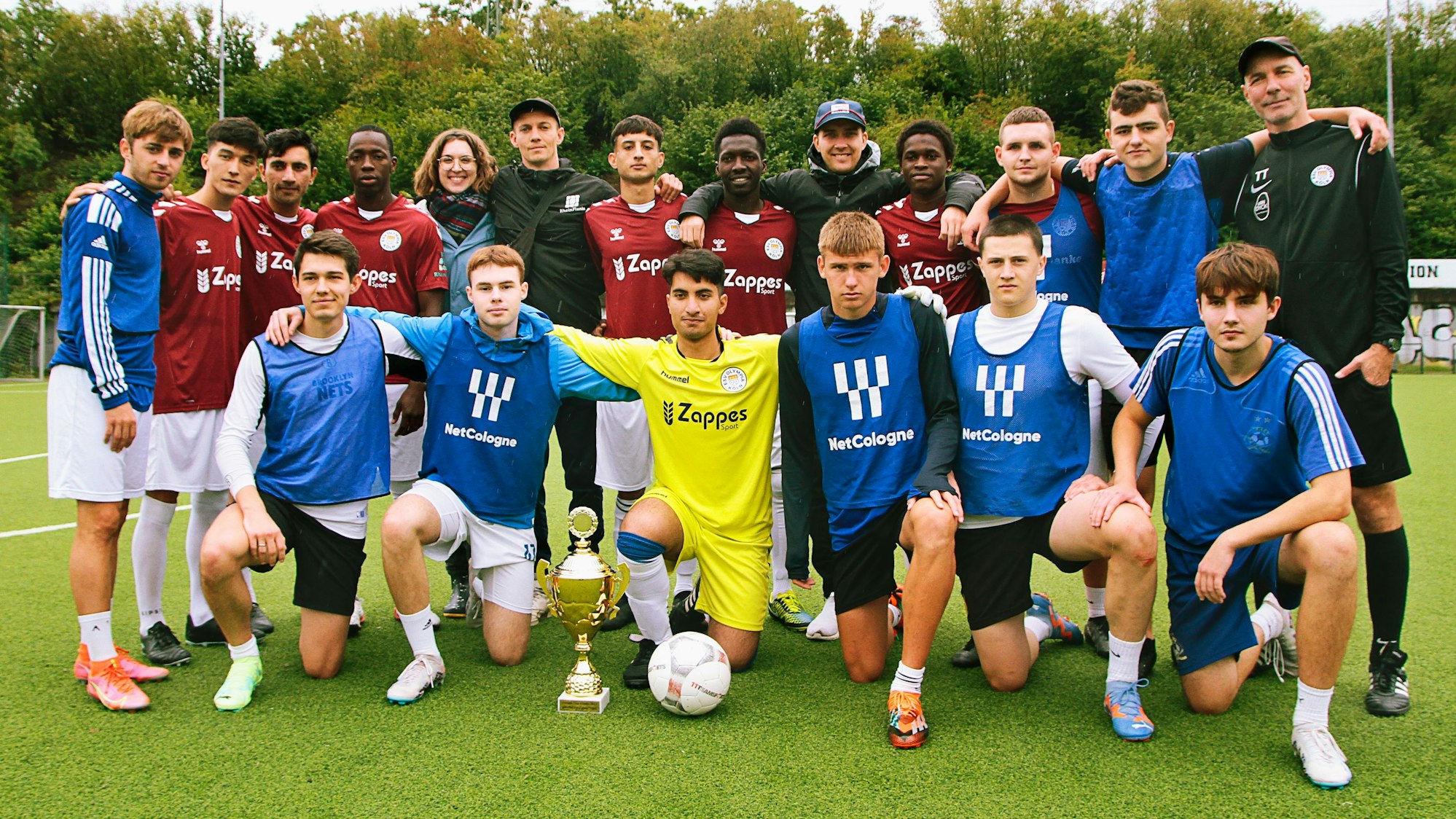 Die Finalisten des von der „Rheinflanke“ organisierten „Nippes-Veedelscup 2023", die „Ukraine Pittbulls“ und „Hope Rheinflanke“ stehen im Nippeser Olympia Stadion, vor ihnen auf dem Boden der Pokal zu Ehren des verstorbenen Fabian S.