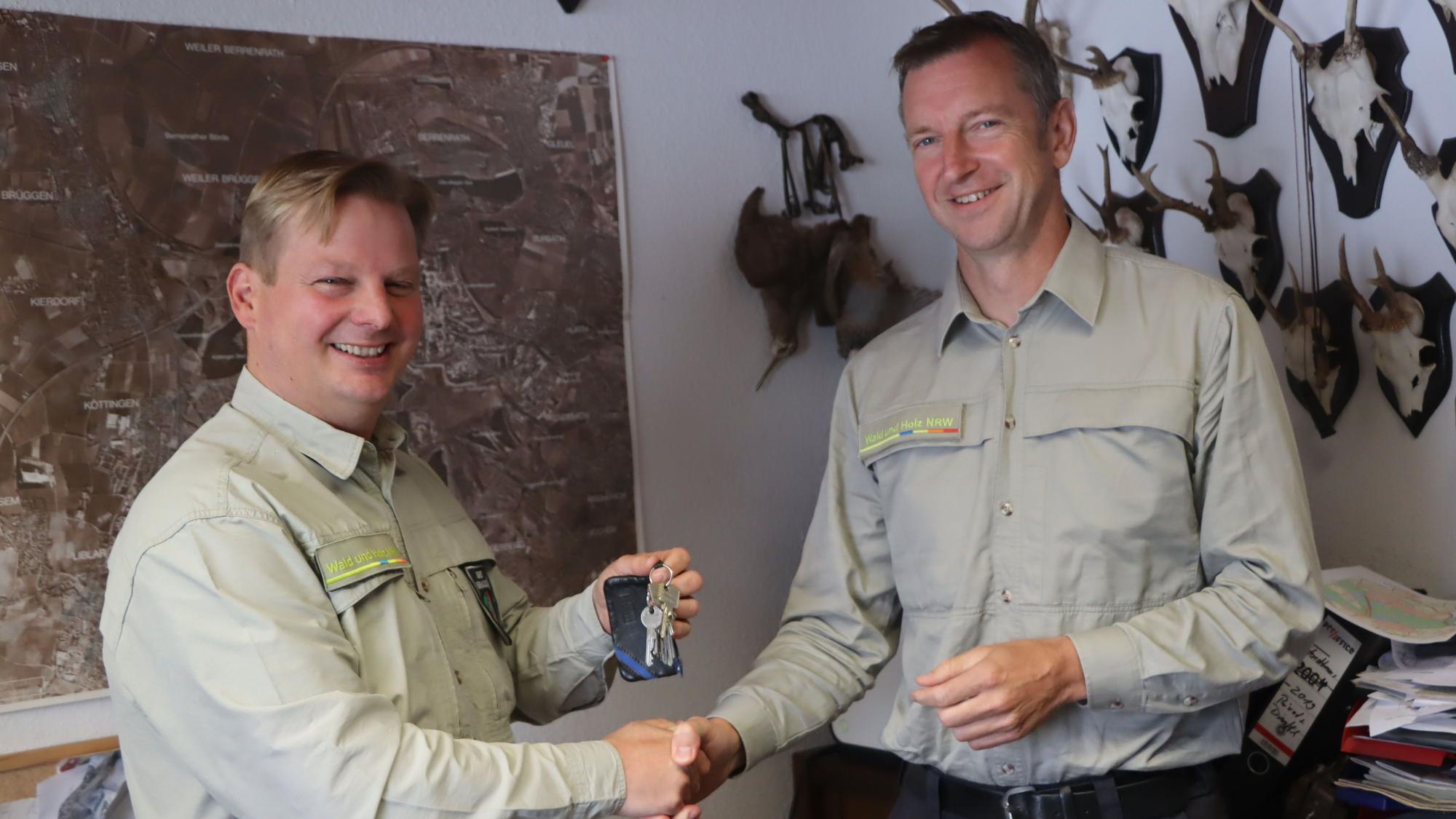 Zwei Männer schütteln sich vor einer Wand mit Landkarte und Jagdtrophäen die Hand.