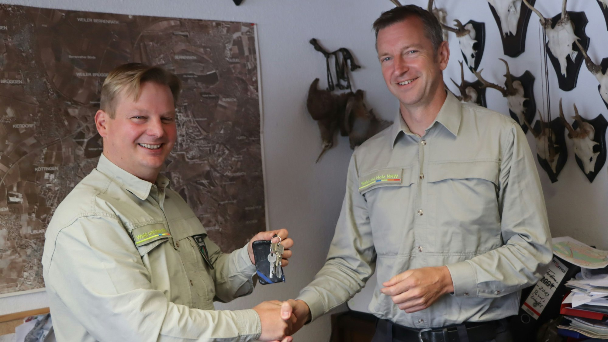 Zwei Männer schütteln sich vor einer Wand mit Landkarte und Jagdtrophäen die Hand.