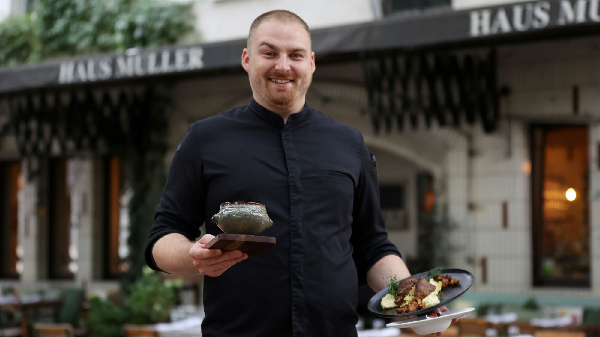 Lasse Hülsmeier steht vor dem Haus Müller und hält drei verschiedene Gerichte in der Hand
