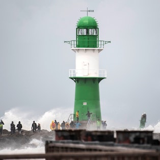 Schwerer Wellengang durch das Sturmtief „Zacharias“ an der Ostseeküste. Mehrere Schaulustige beobachten, wie die Wellen neben einem Leuchtturm bei Warnemünde an die Küste klatschen.