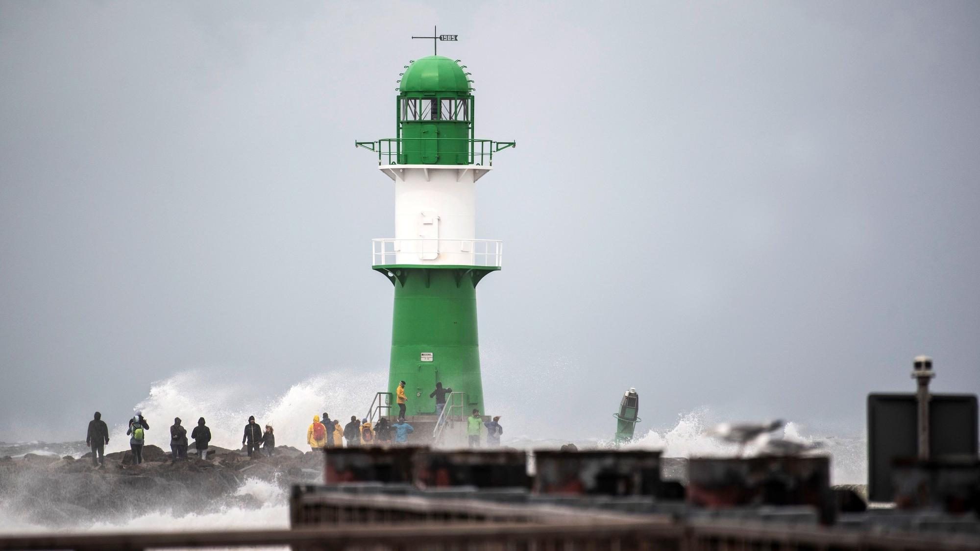 Schwerer Wellengang durch das Sturmtief „Zacharias“ an der Ostseeküste. Mehrere Schaulustige beobachten, wie die Wellen neben einem Leuchtturm bei Warnemünde an die Küste klatschen.