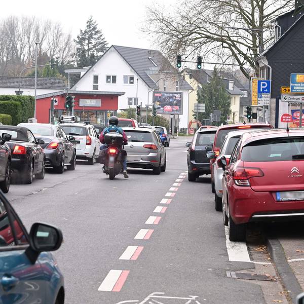 Zu sehen sind zwei Fahrspuren mit einem Rückstau vor der Ampelkreuzung. Der Radstreifen wird durch ein parkendes Auto blockiert. Der Parkstreifen ist vollständig belegt mit parkenden Autos.