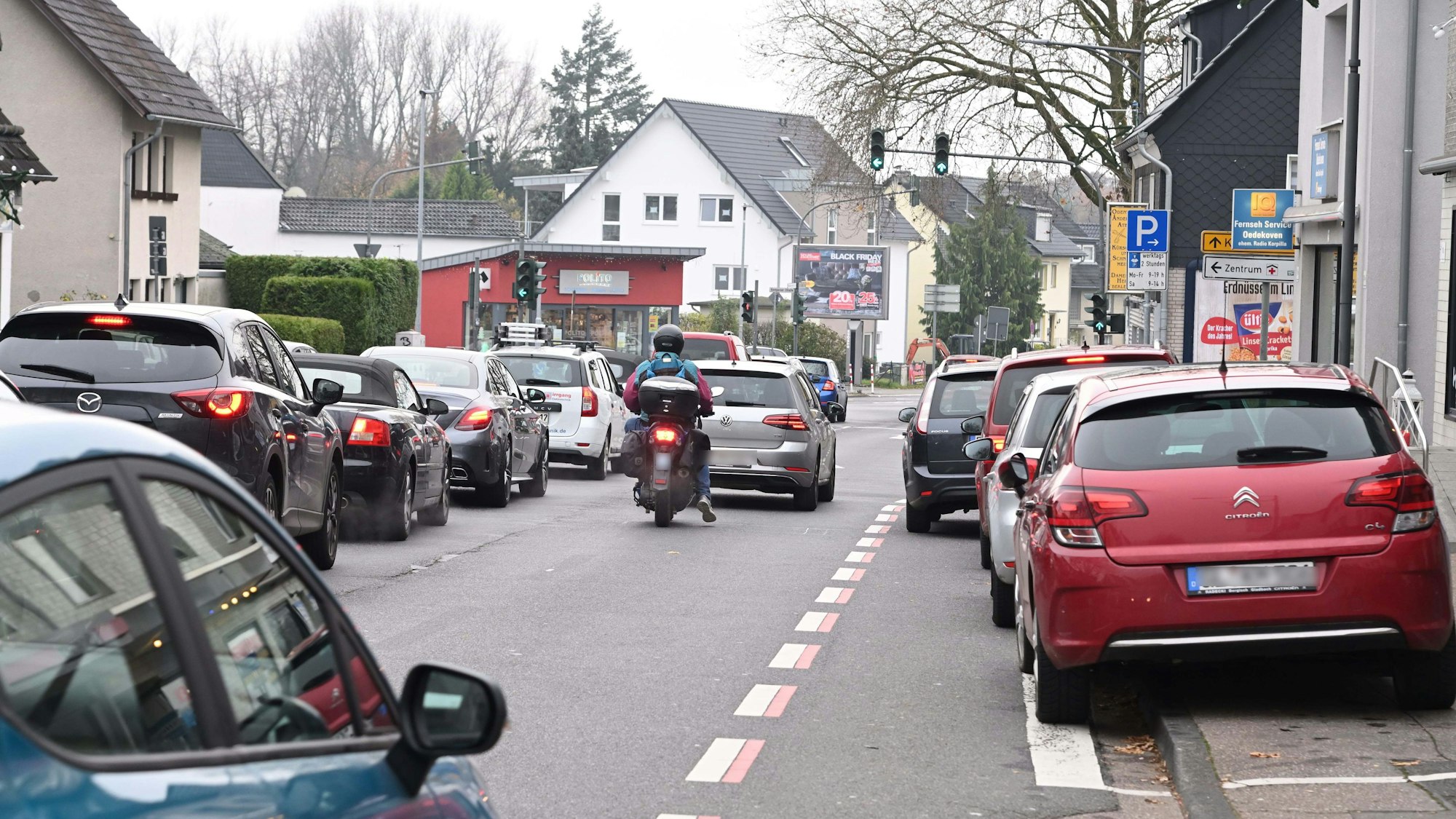 Zu sehen sind zwei Fahrspuren mit einem Rückstau vor der Ampelkreuzung. Der Radstreifen wird durch ein parkendes Auto blockiert. Der Parkstreifen ist vollständig belegt mit parkenden Autos.