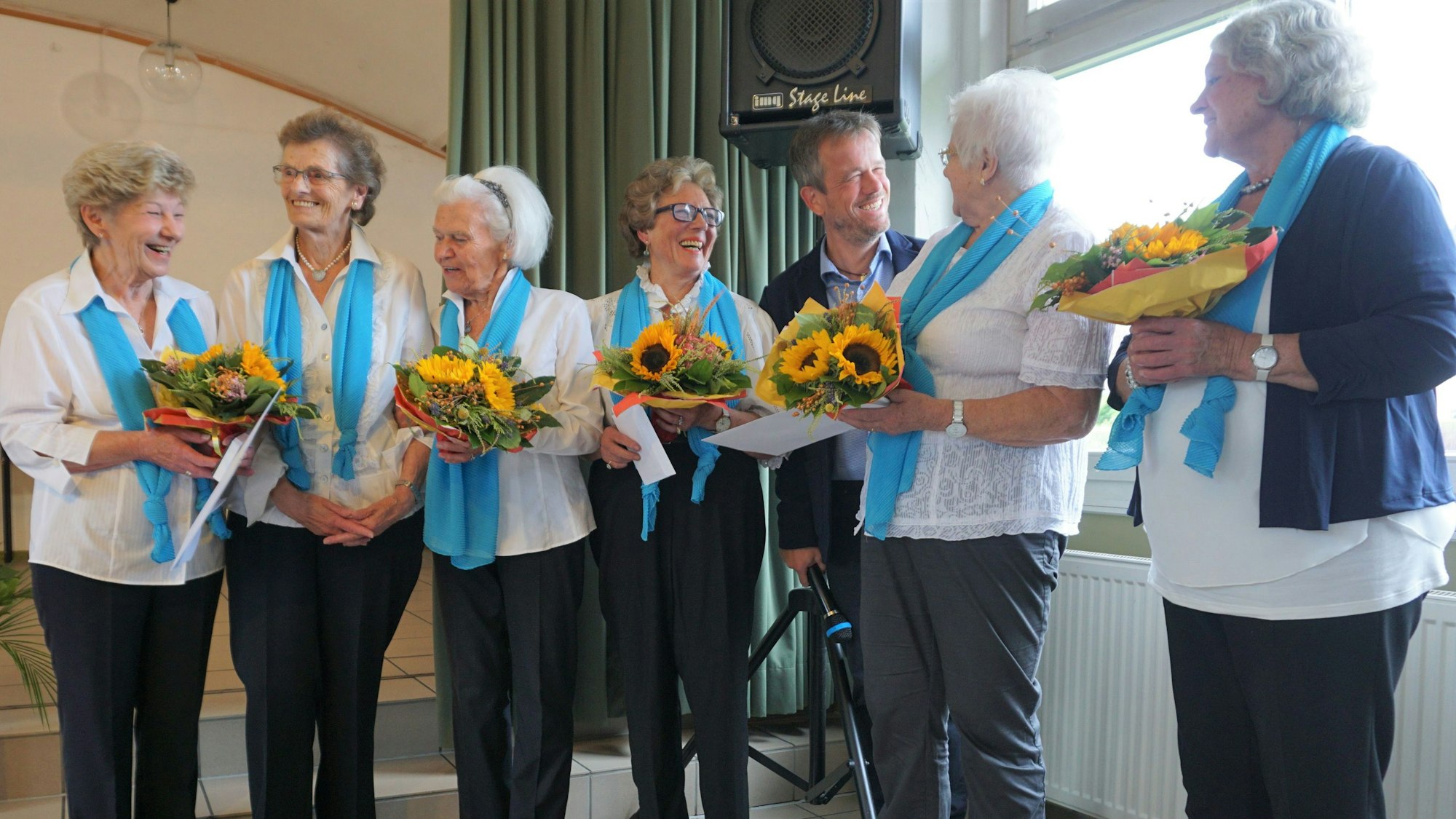 Frauen des Teams in weißen Hemden und hellblauen Schals stehen in einer Reihe. Sie haben Blumensträuße in der Hand.
