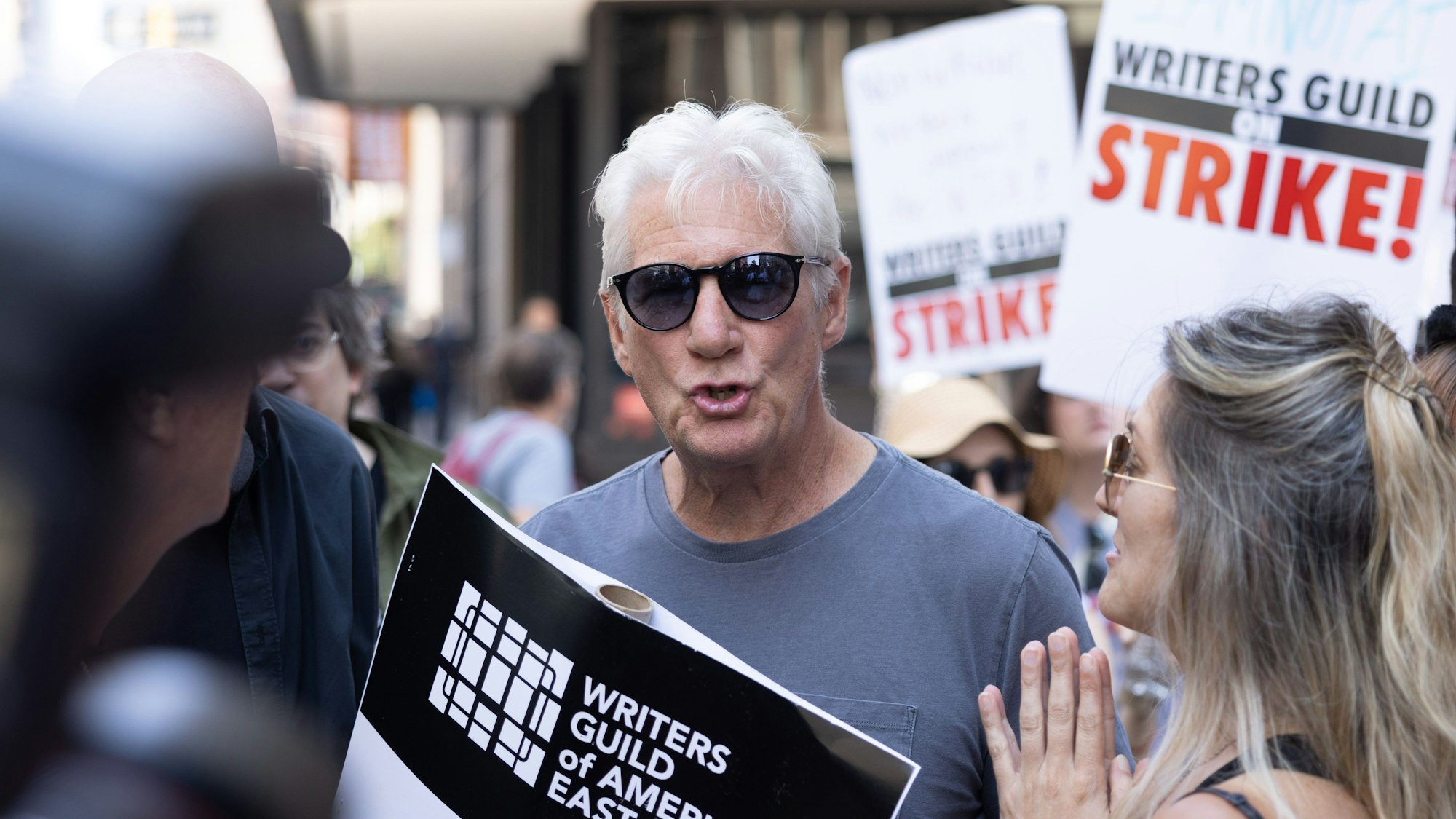 Schauspieler Richard Gere steht mit Sonnenbrille inmitten von streikenden Drehbuchautoren und -autorinnen mit Plaketen.