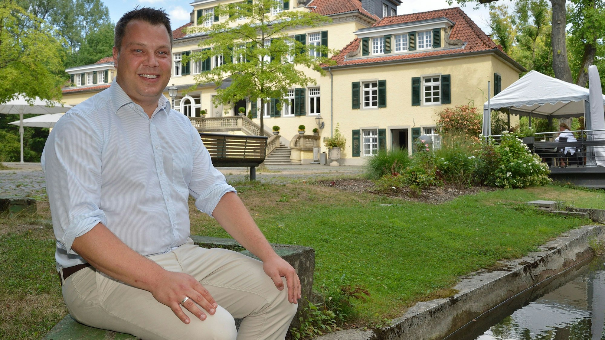 Rösraths Vize-Bürgermeister Sebastian Gopp (FDP) sitzt auf einer Bank vor Schloss Eulenbroich.