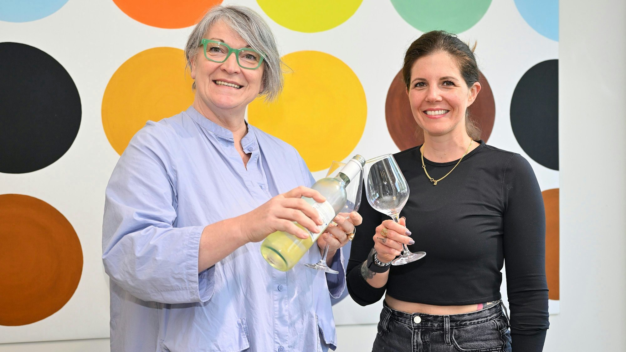 Dagmar Reitz und Katrin Gebauer vom Vorstand der IG Refrath stehen nebeneinander und gießen sich aus einer Weinflasche Wein ins Glas.
