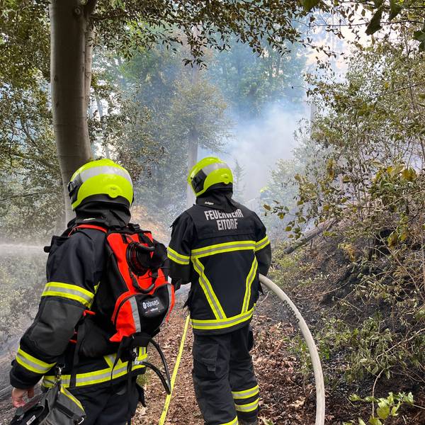 Zwei Einsatzkräfte, die den Schriftzug der Eitorfer Feuerwehr auf dem Rücken tragen, spritzen aus Schläuchen Wasser in den brennenden Wald.
