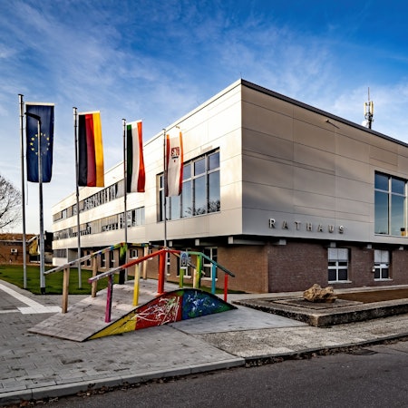 Vor einem großen würfelförmigen Bau wehen Flaggen an hohen Masten und steht eine bunt bemalte hölzerne Brücke.