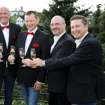 Sechs Menschen stehen auf einer Dachterrasse, stoßen mit Sektgläsern an. Im Hintergrund ist das Bensberger Schloss zu sehen.