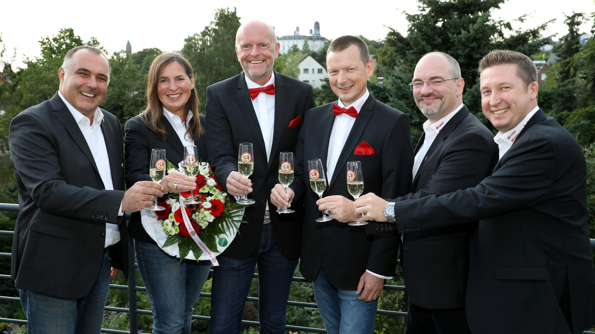 Sechs Menschen stehen auf einer Dachterrasse, stoßen mit Sektgläsern an. Im Hintergrund ist das Bensberger Schloss zu sehen.