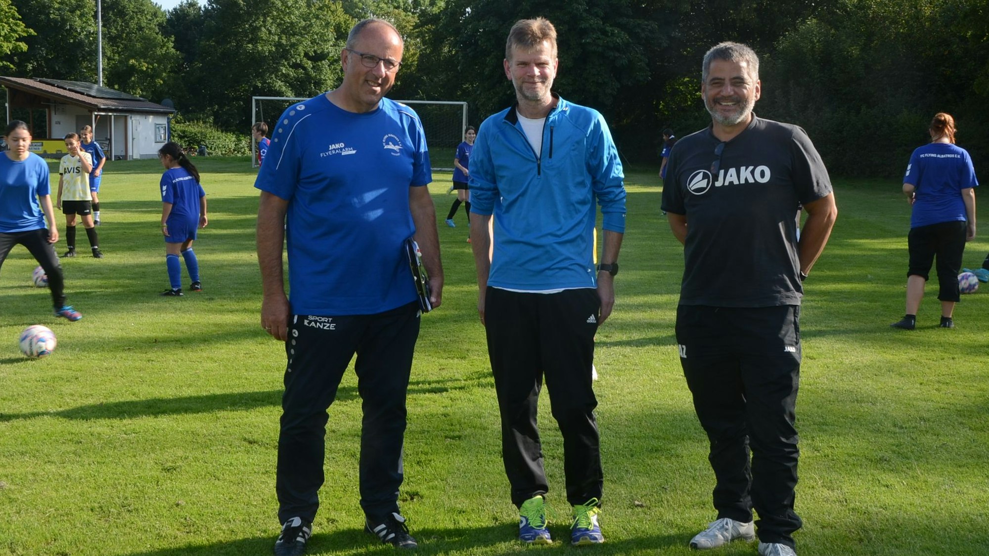Drei Männer in Sportkleidung; im Hintergrund trainieren die Mädchenfußballerinnen.
