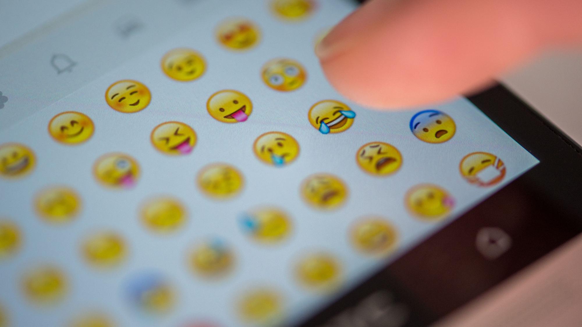 Eine Frau tippt auf das Display eines Smartphones, auf dem zahlreiche Emojis in der Nachrichten-App Whatsapp zu sehen sind.