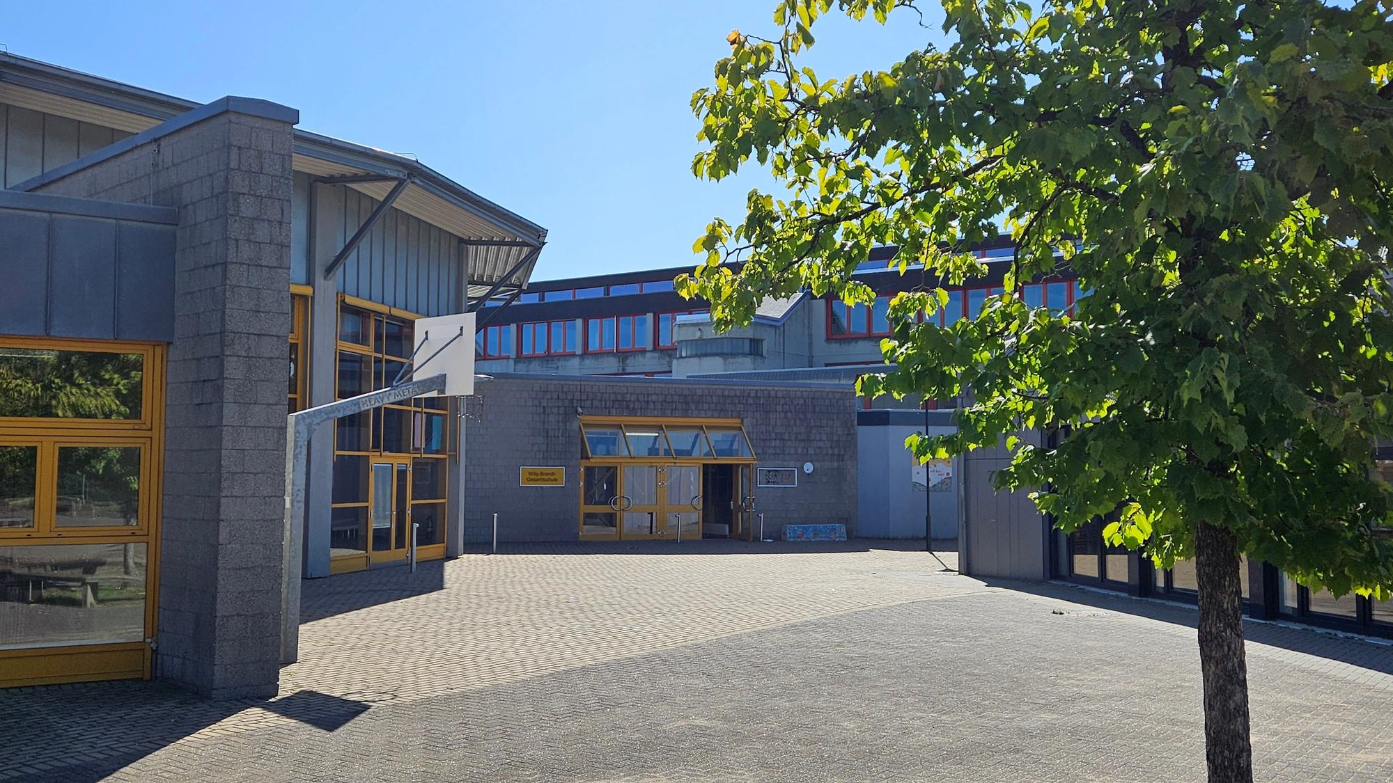 Ein junger Baum steht vor einem modern wirkenden grauen Schulgebäude mit gelben Fenster- und Türrahmen.